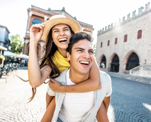 aIm Vordergrund ist ein Pärchen. Er hat sie Huckepack. Beide lachen. Im Hintergrund ist die Szenerie einer kleinen alten Stadt mit Vorplatz zu erkennen.