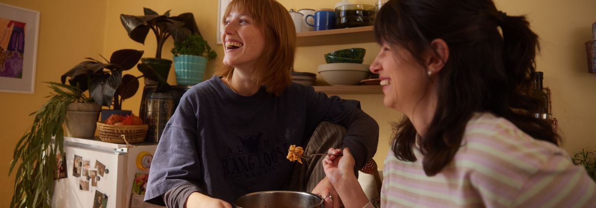 Zwei Freundinnen essen gemeinsam aus einem Topf Nudeln. Die Hintere sitzt auf der Küchenzeile, während sie die Frau im Vordergrund an die Küchenzeile anlehnt.
