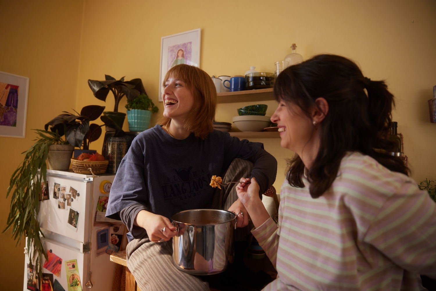 Zwei Freundinnen essen gemeinsam aus einem Topf Nudeln. Die Hintere sitzt auf der Küchenzeile, während sie die Frau im Vordergrund an die Küchenzeile anlehnt.