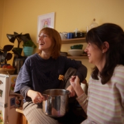 Zwei Freundinnen essen gemeinsam aus einem Topf Nudeln. Die Hintere sitzt auf der Küchenzeile, während sie die Frau im Vordergrund an die Küchenzeile anlehnt.