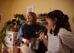 Zwei Freundinnen essen gemeinsam aus einem Topf Nudeln. Die Hintere sitzt auf der Küchenzeile, während sie die Frau im Vordergrund an die Küchenzeile anlehnt.