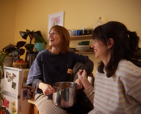 Zwei Freundinnen essen gemeinsam aus einem Topf Nudeln. Die Hintere sitzt auf der Küchenzeile, während sie die Frau im Vordergrund an die Küchenzeile anlehnt.