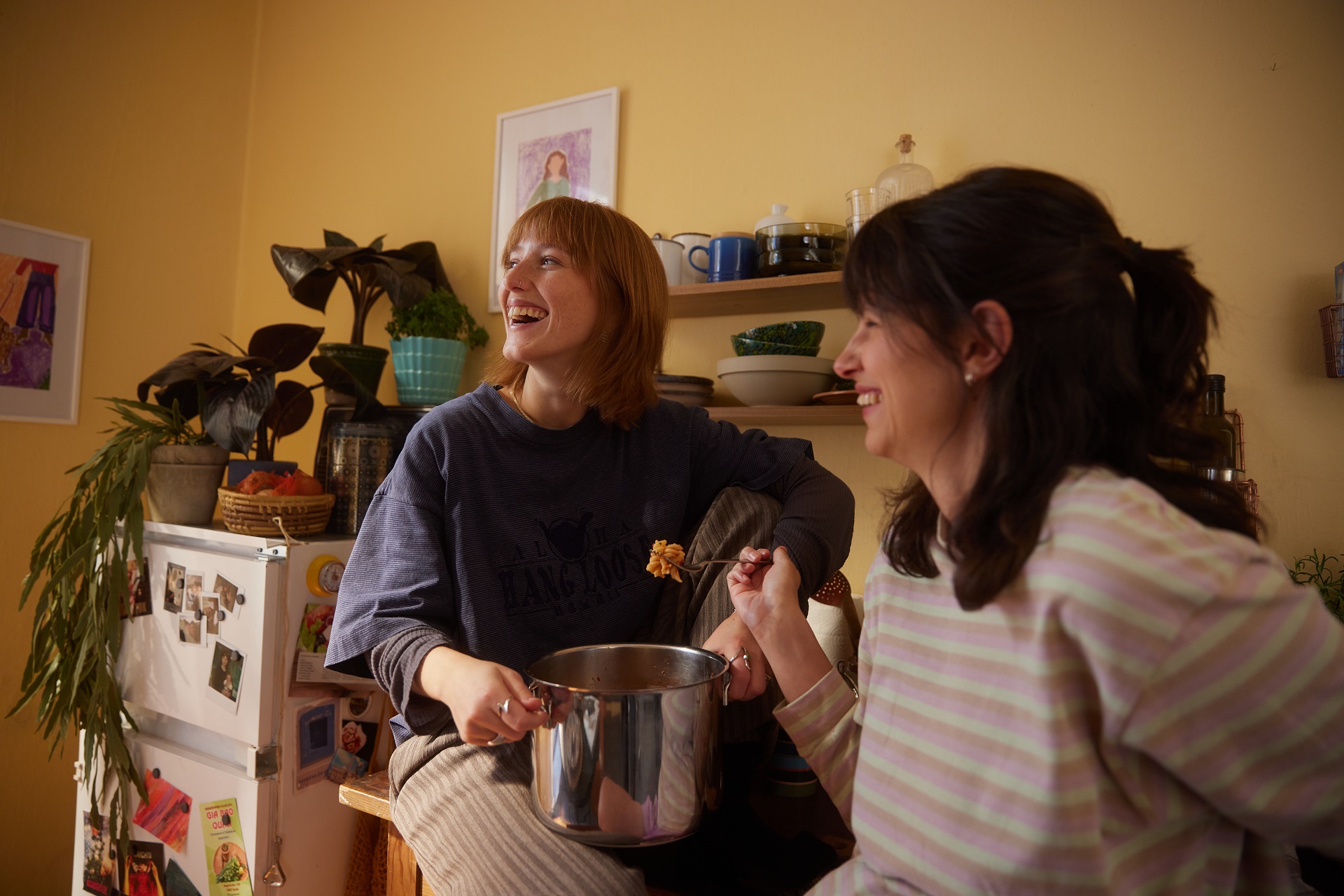 Zwei Freundinnen essen gemeinsam aus einem Topf Nudeln. Die Hintere sitzt auf der Küchenzeile, während sie die Frau im Vordergrund an die Küchenzeile anlehnt. Zwei Freundinnen essen gemeinsam aus einem Topf Nudeln. Die Hintere sitzt auf der Küchenzeile, während sie die Frau im Vordergrund an die Küchenzeile anlehnt.