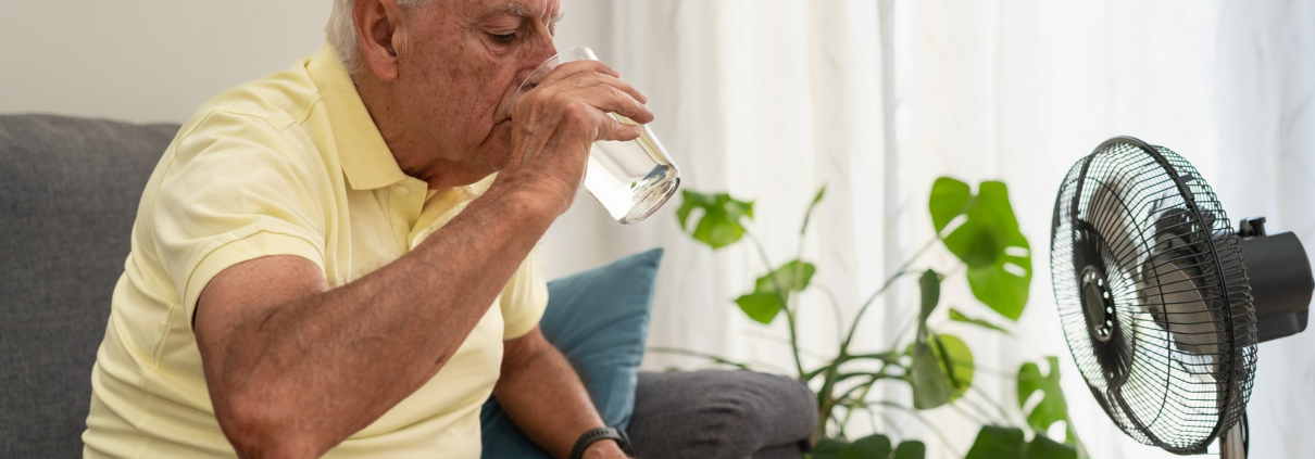 Ein älterer Mann sitzt auf einer Couch und trinkt ein Glas Wasser. Vor ihm steht ein Ventilator.