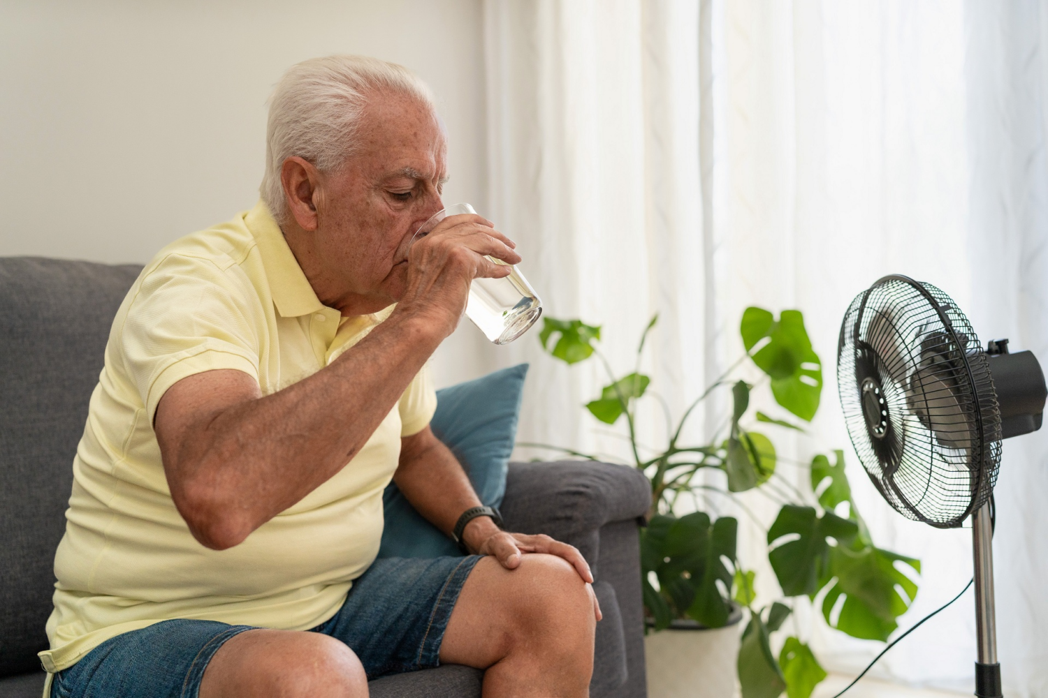 Ein älterer Mann sitzt auf einer Couch und trinkt ein Glas Wasser. Vor ihm steht ein Ventilator.