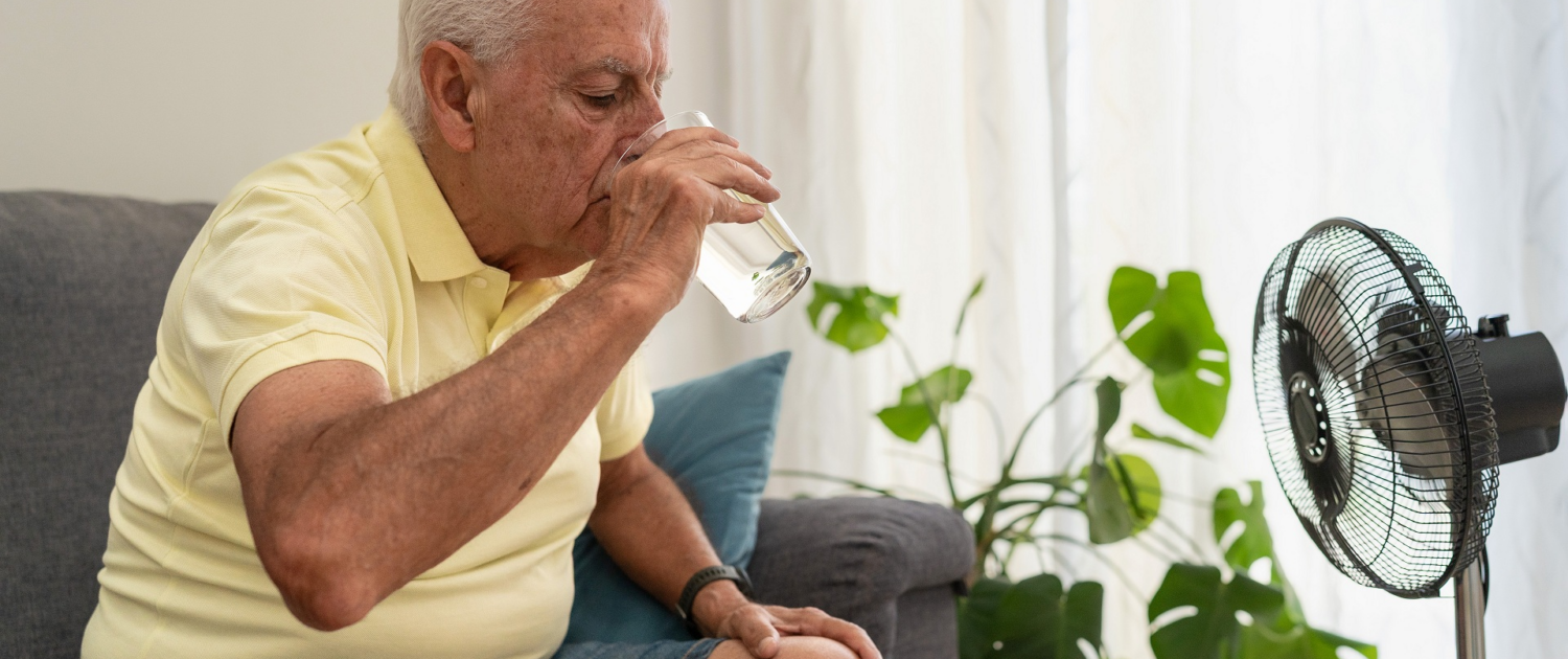 Ein älterer Mann sitzt auf einer Couch und trinkt ein Glas Wasser. Vor ihm steht ein Ventilator.