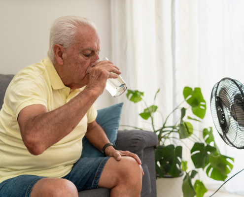 Ein älterer Mann sitzt auf einer Couch und trinkt ein Glas Wasser. Vor ihm steht ein Ventilator.