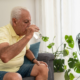 Ein älterer Mann sitzt auf einer Couch und trinkt ein Glas Wasser. Vor ihm steht ein Ventilator.